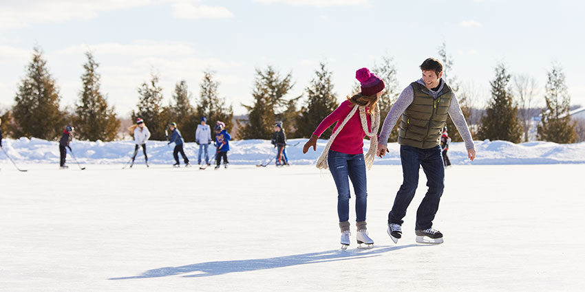 SisterBay-Website-Seasonal-Winter4-850x425_041025 A man and woman skating on an ice skating rink with kids playing hockey in the background in Sister Bay